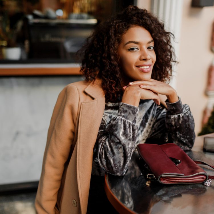 Outdoor  portrait of  beautiful  smiling black woman with stylish afro hairs sitting in cafe in Paris. Wearing trendy velvet dress and beige coat. Luxury bag.